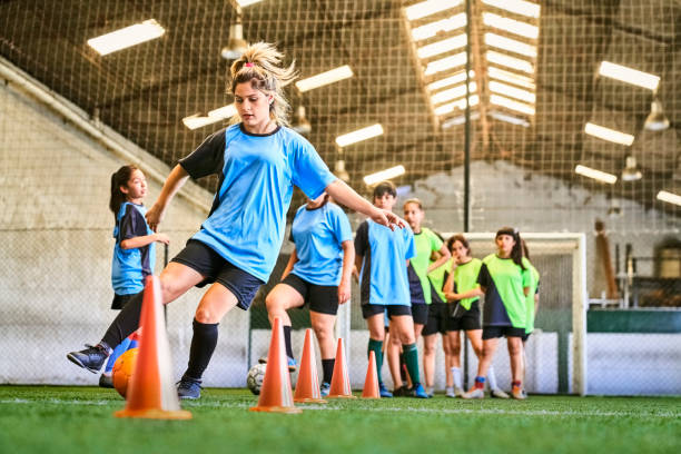 Soccer girl doing dribbling drills on indoor court Group of girls on soccer training at indoor soccer court, doing dribbling drills. Teenage girl in blue jersey kicks soccer ball around orange cones. sports training drill stock pictures, royalty-free photos & images