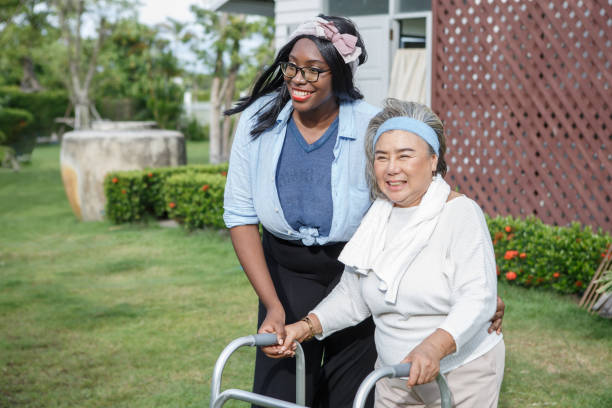 asian senior mother walk with walker and african american carer support in garden at home. old woman disabled walking and black caregiver young woman helping in back yard park outdoors. health care stock photo