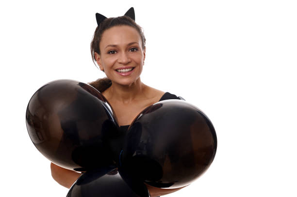 Latin American beautiful woman dressed in black, wearing hoop with cat ears, smiles toothy smile holds black air balls and looks at camera against white background. Halloween, Black Friday Beautiful woman dressed in black, wearing hoop with cat ears, smiles toothy smile holds black air balls and looks at camera against white background. Halloween, Black Friday concept for ad black cat costume stock pictures, royalty-free photos & images