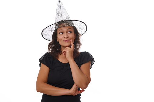 Charming brunette woman in witch carnival hat and black outfit, holds finger on her chin and smiles with a cute toothy smile, staring mysteriously at copy space white background. Halloween concept