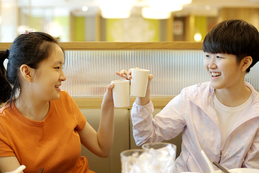 Two Asian teenage girls are holding tea cups for celebratory toast in Chinese restaurant. 

In Chinese culture, the cup that is at higher level represents older age person or better corporate position than the others.