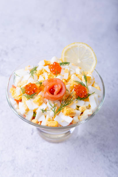 Puff salad with salted trout (salmon), caviar, egg and vegetables. A traditional dish, one serving in a glass glass on a gray background. Croup