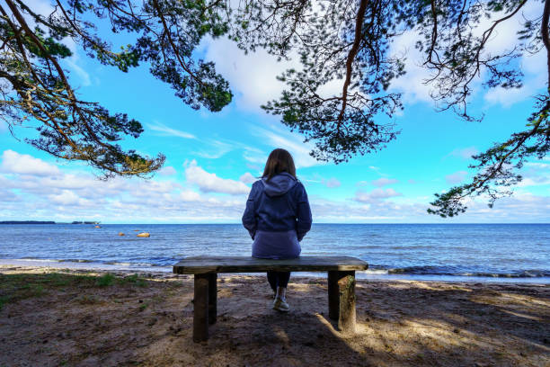 donna da dietro seduta su una panchina di fronte al mare e rami di alberi intorno a lei. - coastline branch day summer foto e immagini stock