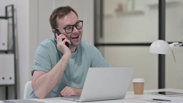 istock Young Man with Laptop Talking on Phone 1347080649
