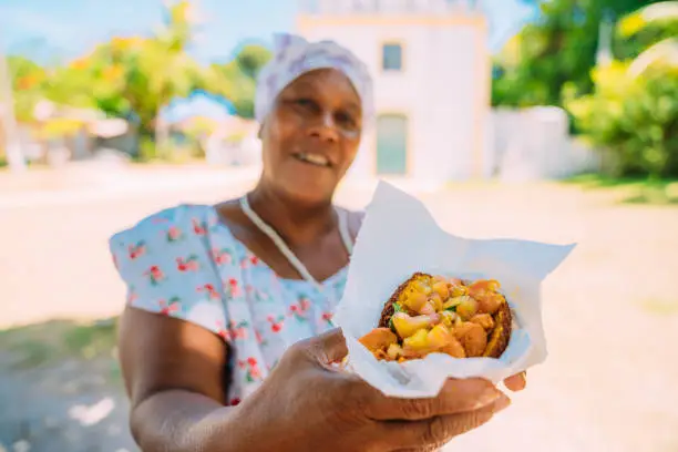 Happy Brazilian woman dressed in the traditional Bahian costume of the Umbanda religion, offering acarajé. Typical Bahia food in the historic center of Porto Seguro in the background.