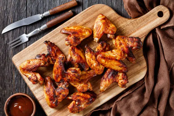 Photo of crispy fried chicken wings served on a wooden board with barbeque sauce on dark wooden table, flat lay