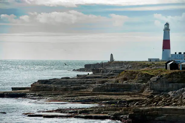 Portland Bill Lighthouse and Obelisk on a rocky shoreline. The Isle of Portland, is a tied island made of the famous Portland Stone in the English Channel. Portland Bill at the southern tip lies south of Weymouth and is the southernmost point of the county of Dorset, England. Chesil Beach a geological shingle barrier beach joins it to the mainland