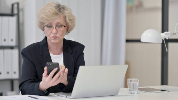 istock Old Businesswoman Browsing Internet on Smartphone while Working on Laptop 1347086145