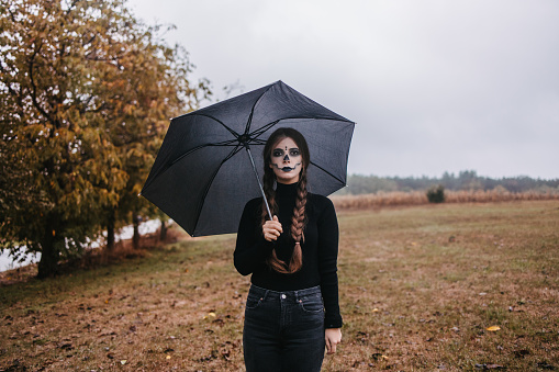 Woman in black with stage make-up and braids holding a black umbrella  on a moody day on a desolate field