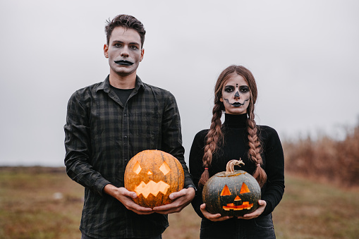 Medium shot of a young couple disguised as zombies, wearing black clothes and with stage make-up holding two curved pumpkins