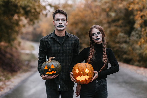 Medium shot of a disguised young couple in black clothes with stage make-up holding two carved pumpkins with candlelight and standing on the road