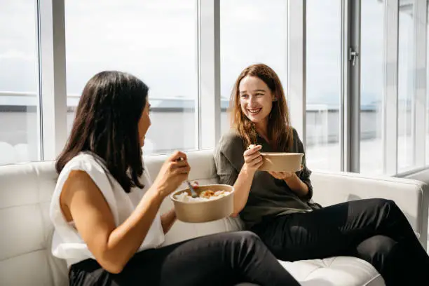 Two female co-workers are sitting on a beige sofa in a bright modern office and enjoying a healthy take-away lunch. The office has large windows. Horizontal daylight indoor photo.