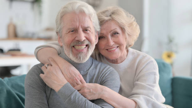 istock Embracing Old Couple Smiling at Camera while Sitting on Sofa 1347092986