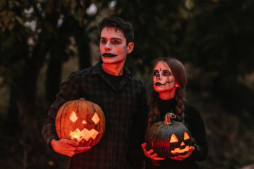Medium shot of a disguised young couple in black clothes with stage make-up holding two carved pumpkins with candlelight and standing on the road looking away illuminated by red light