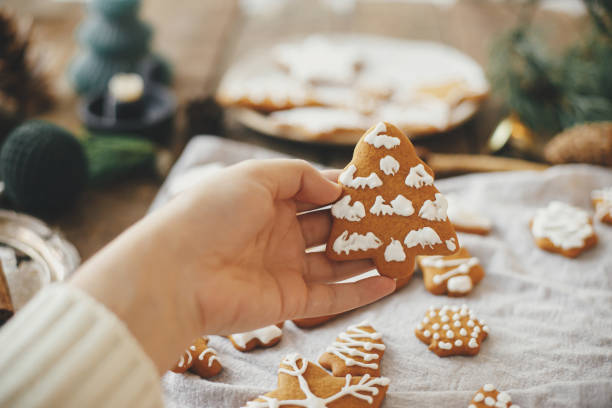 istock Hand holding decorated gingerbread cookie christmas tree on background of rustic table with napkin, candle, decorations. Moody image. Woman making stylish christmas gingerbread cookies 1347098843