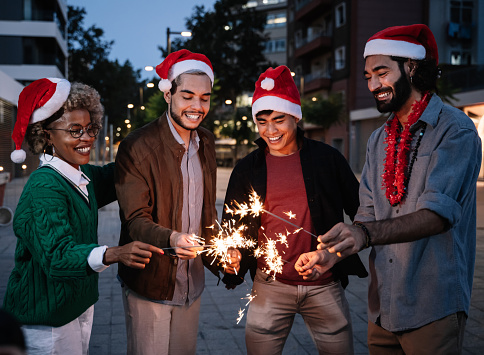 Group of happy multi ethnic friends celebrating Christmas and New Year's Eve with burning sparklers outdoors in the city street after dinner party. Focus on the Asian Man
