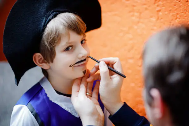 Father making face painting for little kid boy. Child dress up for carnival, halloween or birthday party. Dad doing make up, drawing musketeer hero for son