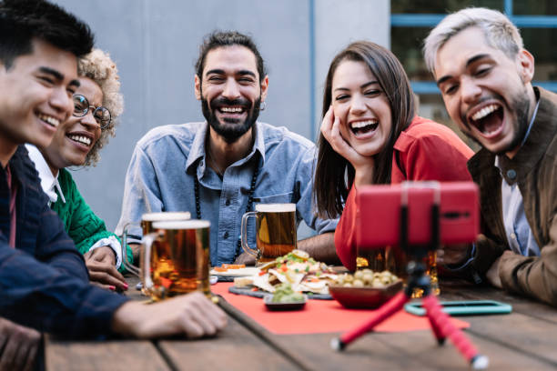 grupo de amigos multirraciais desfrutando comida e bebendo cerveja enquanto faz video-chamada online com celular no bar da cervejaria para comemorar aniversário - foco no rosto do homem - mobile phone group of people human face latin american and hispanic ethnicity - fotografias e filmes do acervo