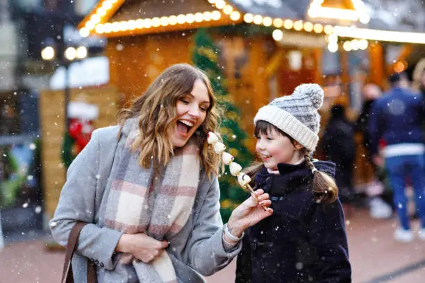 Photo of Mother and daughter eating white chocolate covered fruits and strawberry on skewer on traditional German Christmas market. Happy girl and woman on traditional family market in Germany during snowy day
