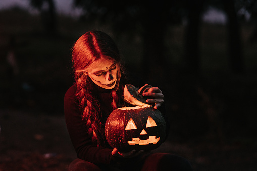 Portrait of a young woman with stage make-up illuminated by red stop lights holding carved Jack o’lantern pumpkin and looking it