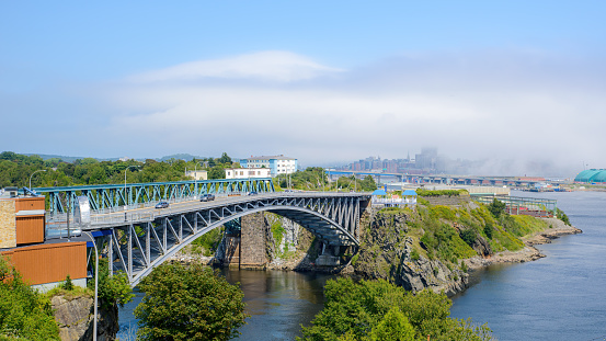 Reversing Falls bridge in Saint John, NB, Canada, seen from Wolastoq Park. Fog creeps over the city center in the distance. Sunny day.