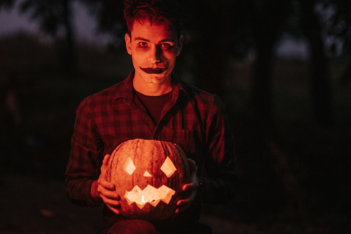 Portrait of a young man with stage make-up illuminated by red stop lights holding curved Jack o’lantern pumpkin and looking at camera