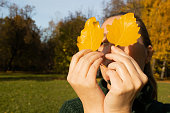Portrait of woman with green eyes eyes holds two yellow leaves, hands close-up, background autumn trees