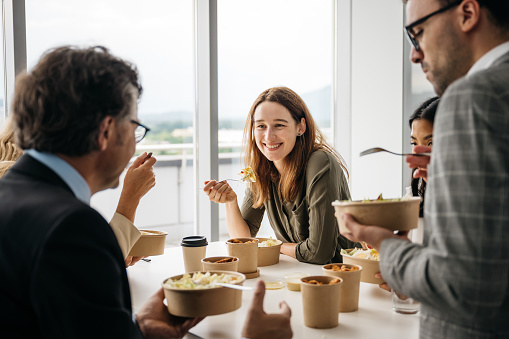 A smiling woman is enjoying a healthy takeaway lunch with her colleagues in a bright modern office. They are laughing and having relaxed chat. Horizontal daylight indoor photo.