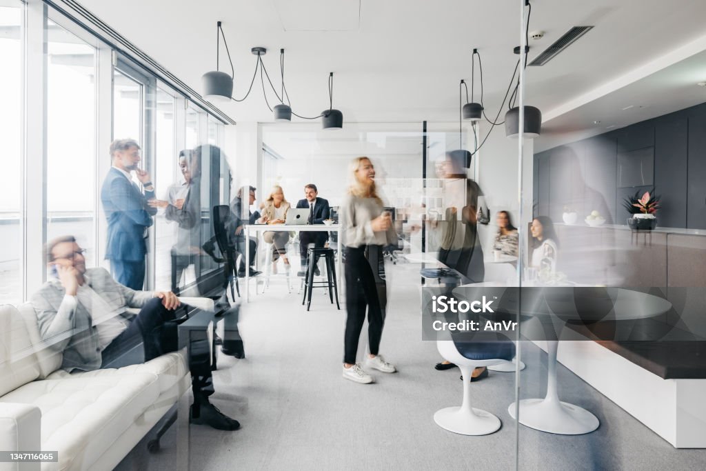 Daily routine at the office A photo showing a daily routine at the office. Some are standing up having conversations, some are sitting down chatting. They are smartly dressed. The office is spacious, modern and has large windows. Horizontal daylight indoor photo. Office Stock Photo