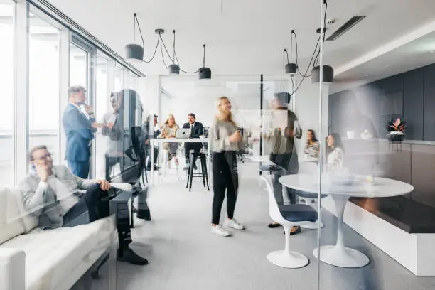 A photo showing a daily routine at the office. Some are standing up having conversations, some are sitting down chatting. They are smartly dressed. The office is spacious, modern and has large windows. Horizontal daylight indoor photo.