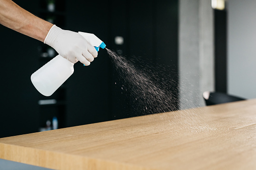 A close-up shot of disinfecting a desk in an office in order to prevent the spread of coronavirus. Only a hand wearing a medical glove and using a disinfectant is visible. Horizontal daylight indoor photo.