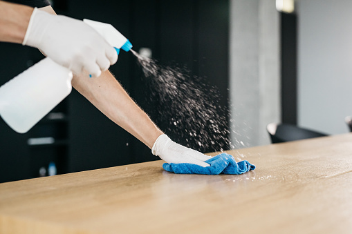 A close-up shot of cleaning a wooden desk in an office with a disinfectant to limit the spread of COVID-19. Only hands wearing medical gloves are visible, no faces. Horizontal daylight indoor photo.