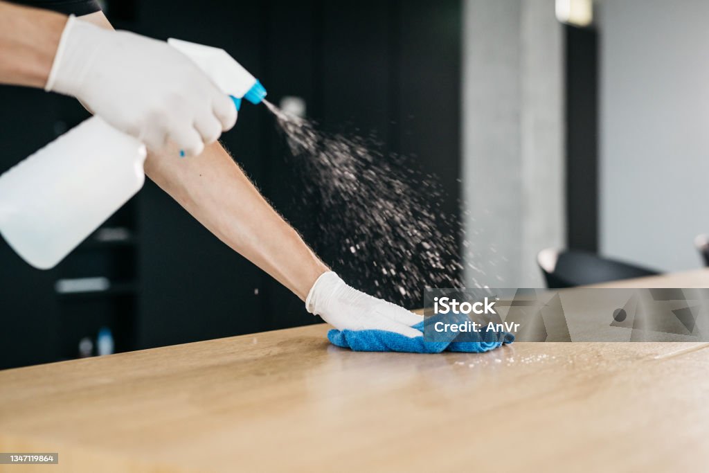 Close-up of disinfecting a wooden desk to limit the spread of COVID-19 A close-up shot of cleaning a wooden desk in an office with a disinfectant to limit the spread of COVID-19. Only hands wearing medical gloves are visible, no faces. Horizontal daylight indoor photo. Cleaning Stock Photo
