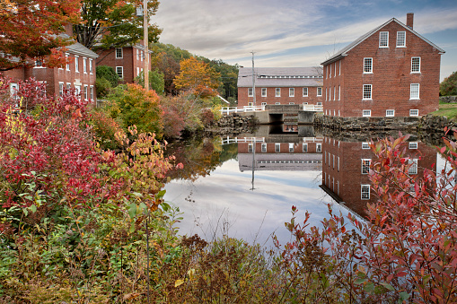 Colorful autumn scene at historic 19th-century textile mill town in Monadnock region of New Hampshire. Picturesque village of Harrisville with reflection of sky and red brick buildings on water.