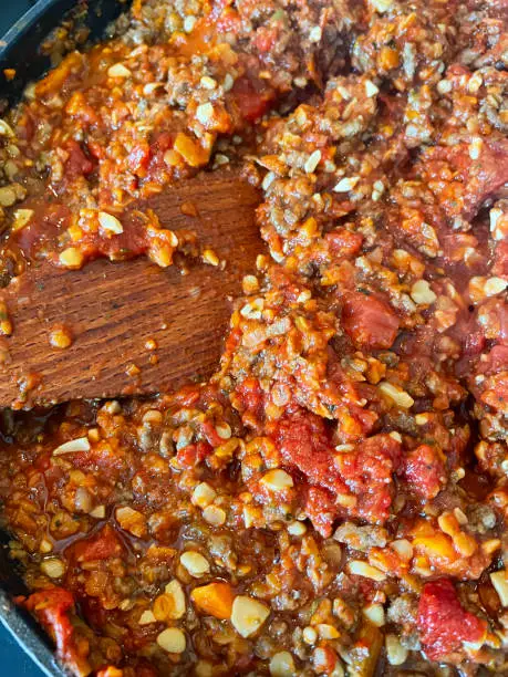 Photo of Full frame image of non stick frying pan containing partially cooked lean minced beef, minced meat browning with finely chopped carrots, onions, walnuts and red lentils, preparing bolognese sauce, stirring with wooden spatula, elevated view
