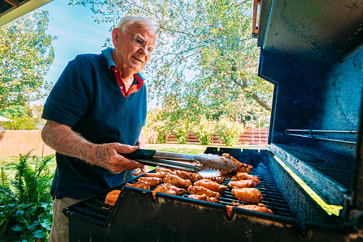 Senior Caucasian Man Standing Outdoors on the Back Patio Checking the Doneness of Spicy, Delicious Buffalo Chicken Wings on the Grill for a Summer Afternoon Lunch or Dinner Party