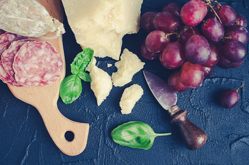 Assorted Italian style food set - salami, Parmesan cheese, grapes and basil. Mediterranean appetizers, tapas or antipasti on black background. Delicious snack on party. Top view.