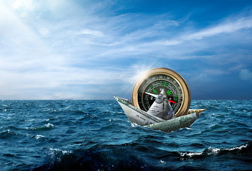 Conceptual money boat carrying large compass in stormy seascape