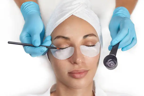 Photo of Close-up of the girl's face with closed eyes. The hand of the master tint to the eyelash with a cotton swab in cosmetology clinic.