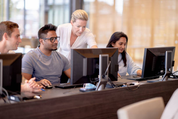 Multi-Ethnic Mature Students in a Computer Lab A group of mature students each sit in front of a desktop in a computer lab.  They are each dressed casually and focused on their screens as their female teacher makes her way around to check on each student individually. computer lab stock pictures, royalty-free photos & images