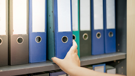 Female hand holding a binder of documents on the row of file folders that nicely management system on the office's shelves.