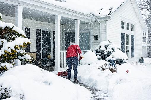 A senior adult woman wearing a warm red winter jacket coat is using a manual snow shovel to push and clear away the pedestrian walkway footpath in front of her house of deep winter snow while a western New York State February blizzard storm continues dropping more in this suburban residential district near Rochester, New York.