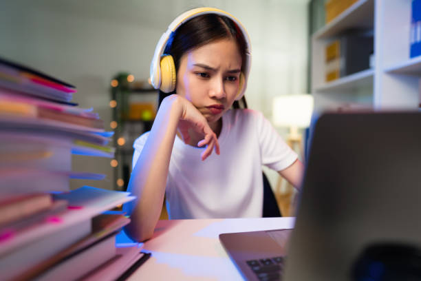 Stressful  young Asian woman wearing headset and sitting on the chair with a pile of papers document on the table and looking on the computer screen on night. stock photo