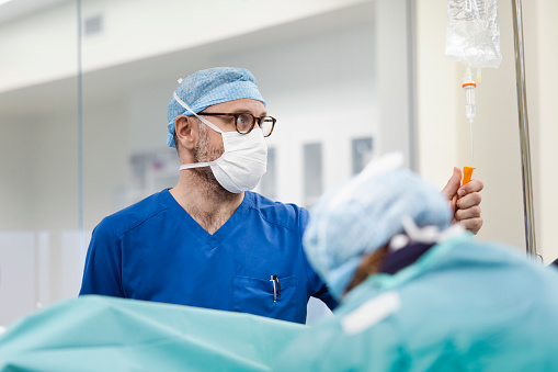 Male anesthetist adjusting IV drip in operating room. Medical professional is working in operating room. He is wearing protective face mask.