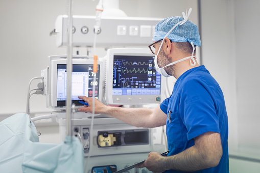Anesthetist examining monitoring equipment in operating room. Male medical professional is working at hospital. He is wearing surgical cap and face mask.