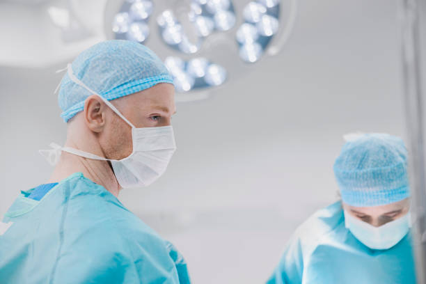 Male and female surgeons doing surgery at hospital Male surgeon looking away during surgery in operating room. Team of doctors working in hospital. they are wearing surgical caps and face masks. surgical light stock pictures, royalty-free photos & images