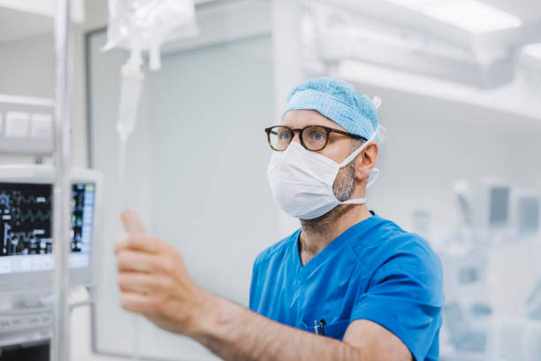 Doctor examining IV drip in operating room Male anesthetist holding IV drip in operating room. Healthcare worker is examining intravenous bag. He is wearing surgical cap and mask. anaesthetist stock pictures, royalty-free photos & images