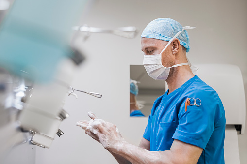 Male surgeon in uniform washing hands in hospital. Male doctor is scrubbing fingers. He is wearing face mask and surgical cap.