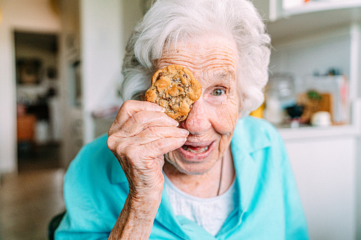 Cute Portrait of a Cheerful, Sharp 100-Year-Old Senior Woman Holding a Sweet Cookie in Front of Her Eye and Making a Face Smiling at the Camera