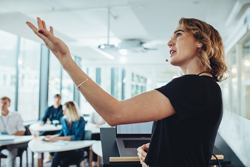 Businesswoman gesturing at projection screen and making a presentation to her colleagues in office. Female speaker giving presentation in conference.